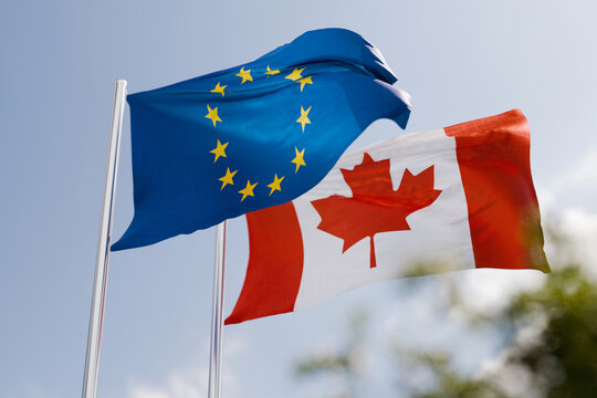 Flags of the European Union and Canada waving in the wind against a blue sky, symbolizing international relations, diplomatic alliance, economic partnership, trade agreements, and cooperation.