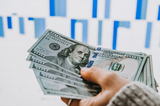 Hands holding US dollars currency in front of the stock exchange screen showing trading graphics.
