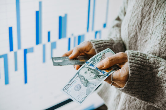 Hands holding US dollars currency and a bank card in front of the stock exchange screen showing trading graphics.