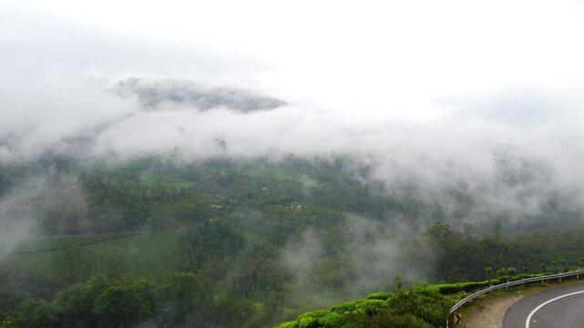 Aerial View of Mountain Pass Highway Corner with Heavy Mist Rolling Over Western Ghats Forests in Keralam. Cloud and Low Mist Passing Across Mountain Range Along Highway Viewpoint.
