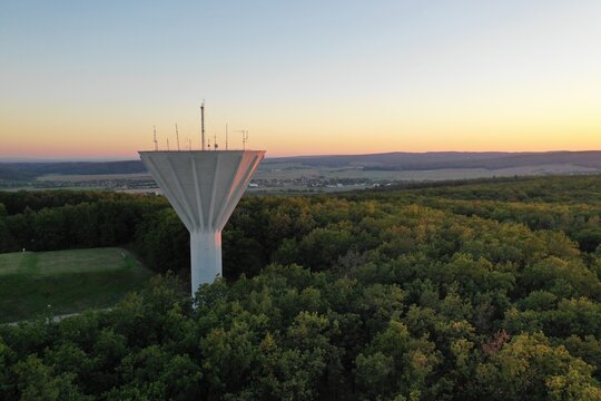 Aerial drone photo of concrete water tower with conical funnel-shaped tank and telecommunication antennas rising above dense green forest canopy at sunset, rural countryside infrastructure photo