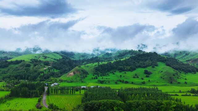 Mist drifts over vibrant rolling hills, patchwork farmland and shelterbelt trees in a tranquil Xinjiang valley beneath dramatic clouds.