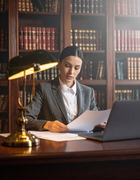 A professional female lawyer studies legal documents in a dimly lit, atmospheric library office. This elegant scene evokes focus, authority, and scholarly dedication to the practice of law.