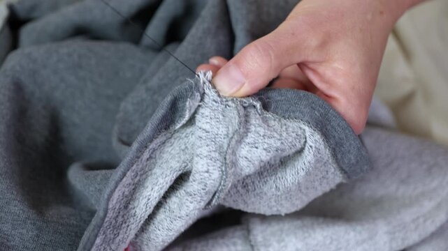 Person hand sewing gray fabric with needle and thread. Close-up view of a skilled tailor's hands meticulously mending a hole in a gray sweatshirt using a traditional needle black thread, and a thimble