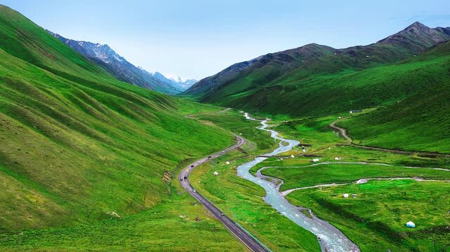 Aerial view of a winding river and highway crossing a vivid green mountain valley beneath rugged snowcapped peaks in Xinjiang.