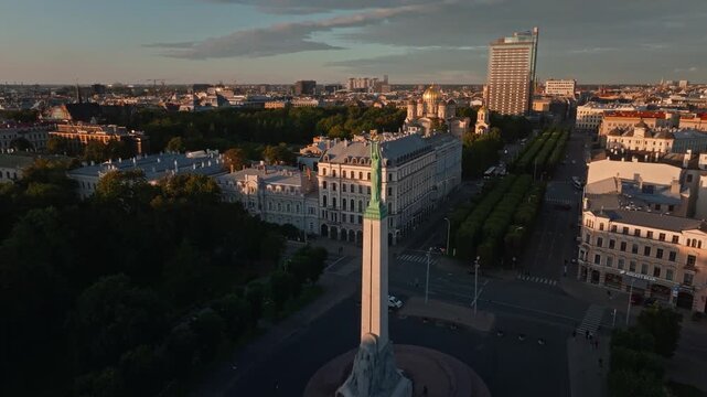 Freedom Monument Milda over boulevard and hotel tower from drone, Riga. Aerial shot orbiting down to the Liberty statue in Riga, Latvia, above tree lined avenue, historic facades.