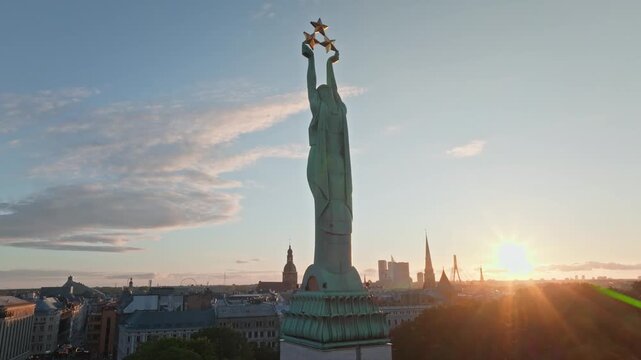 Freedom Monument Milda and old town spires at sunset from drone, Riga. Aerial shot circling the Liberty statue with three stars in Riga, Latvia, under evening clouds above boulevard.
