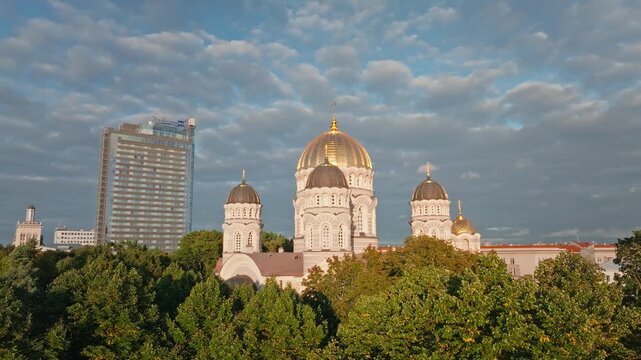 Cathedral golden domes revealed above park trees from rising drone, Riga. Aerial shot climbing over green treetops in Riga, Latvia.