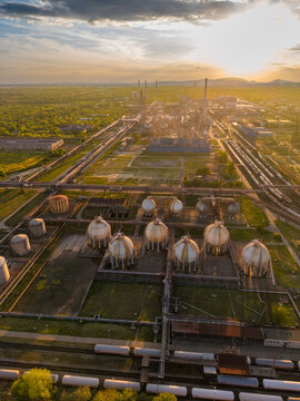 Vertical aerial view of spherical refinery tanks at sunset. White vessels fill the lower half between pipe corridors leading toward stacks beneath a clouded golden sky.