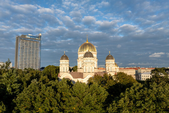 Nativity of Christ Cathedral and hotel tower under evening clouds from drone, Riga. Aerial view of the golden domed orthodox church above green treetops beside the tall hotel tower.