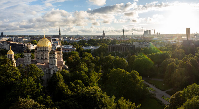 Orthodox cathedral in park with sun flare, drone panorama, Riga, Latvia. Drone wide panoramic view of the golden domed church among park trees with low sun flare, old town spires.