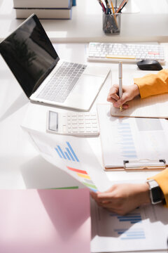 Desk Work Analysis: Close-up shot of a person's hands as they analyze financial charts and graphs on paper, with a laptop and calculator on the desk.