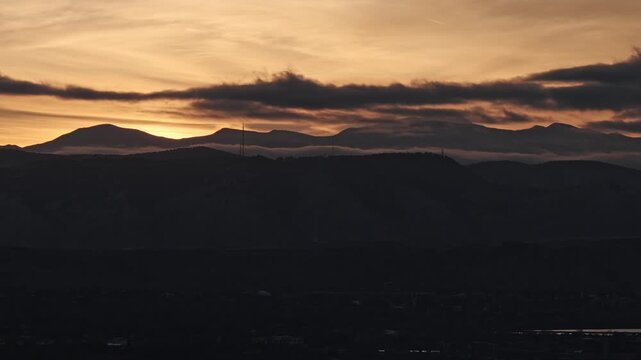 Majestic sunset with golden light disappearing behind a dark mountain silhouette and moving clouds