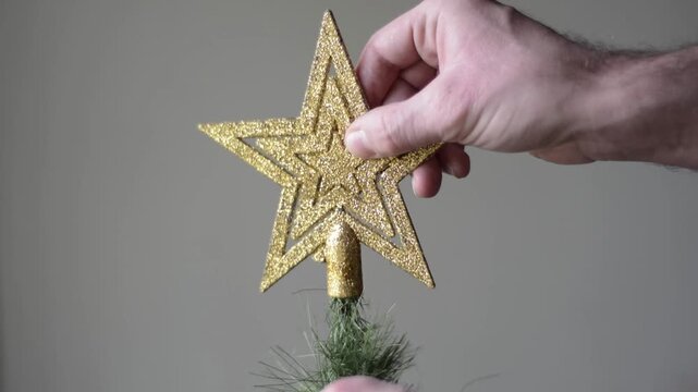 Close up of man's hand placing gold star on top of Christmas tree against flat neutral background