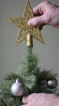 Close up of male hand placing golden star on top of Christmas tree decorated with silver balls against flat grey wall