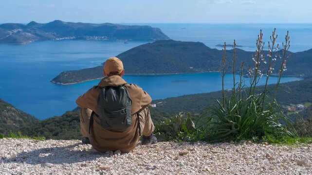 Hiker enjoying the stunning seascape from a mountain top. Young backpacker sitting on a cliff edge, peacefully enjoying a breathtaking panoramic view of the turquoise sea and islands, finding serenity