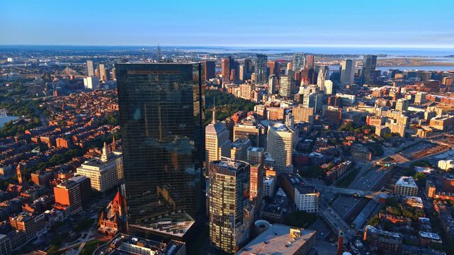 John Hancock Tower above Boston with downtown and harbor in the distance. A dark glass facade dominates the foreground beside a curved tower and parallel road and rail routes.