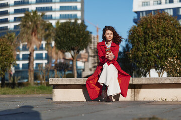 Fototapeta premium A young woman with a red coat sits on a city bench in daylight, looking confident and relaxed in an urban park setting with modern buildings behind, hinting at everyday street style.