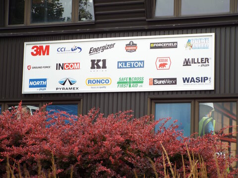 Ottawa, Ontario, Canada &ndash; July 19, 2026: A sign displaying "multiple safety equipment brand logos" including "3M, Energizer, Kleton, Ronco, and Wasip" above red bushes on Canotek Rd.