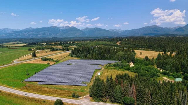 Solar farm between a rural road and the Tatra Mountains. An array of blue modules occupies a clearing beside fields, with layered ridges across the horizon.