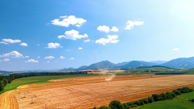 Grain harvest across farmland below mountain ridges. Farm machinery raises a dust plume above a golden field bordered by green plots and distant hills.