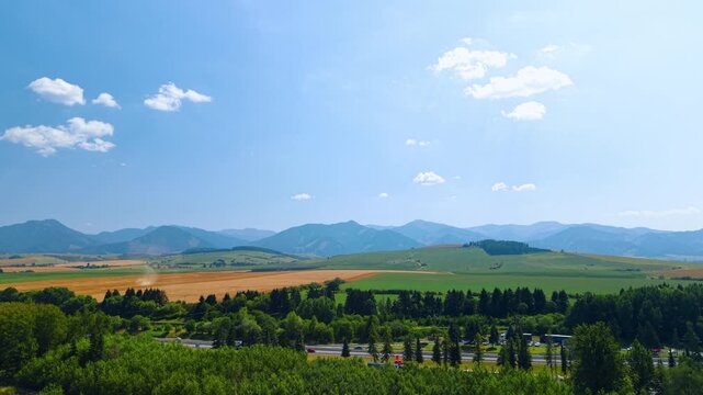 Agricultural valley beyond a highway and woodland. Trees frame divided lanes below green and ochre fields, with a hazy mountain ridge beneath open sky.