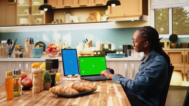 Female worker uses vertical blue screen and chroma key on tablet and laptop while working from home, multitasking with gadgets in modern indoor lifestyle.