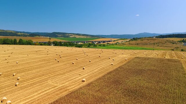 Harvested field and standing crops beside a viaduct. Straw bales dot yellow stubble beside a brown crop strip, with bridge arches below wooded hills.