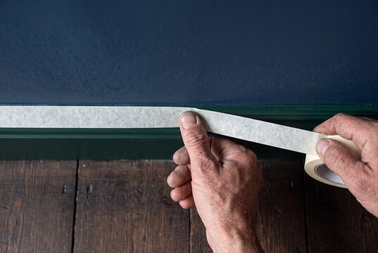 Close-up of a worker applying protective masking tape along a wooden baseboard before painting the interior wall