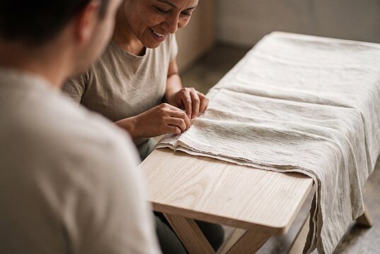 Smiling female artisan working with natural linen fabric on a wooden table in a creative textile workshop with her partner