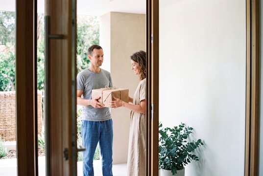 Smiling young woman receiving a wrapped parcel delivery package from a courier at the open doorway of her modern home
