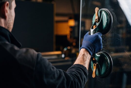 Glazier carrying a large sheet of clear glass using a double suction cup tool in a workshop