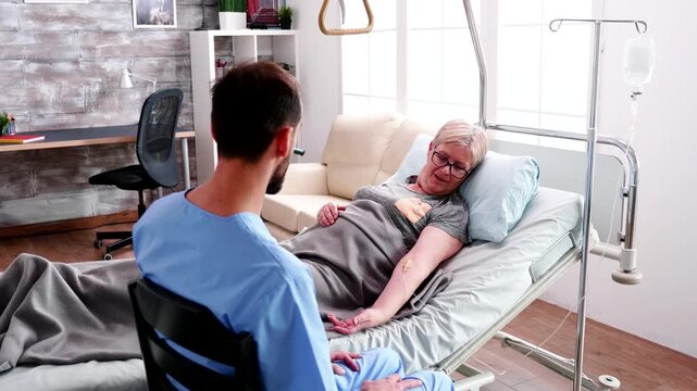 Male healthcare worker sits close to a senior woman in bed as they discuss treatment and care, combining personal communication with supportive elder services.