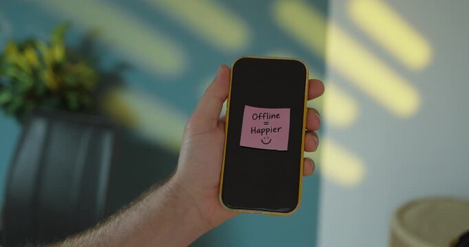 Person places an &ldquo;Offline = Happier&rdquo; sticky note on smartphone, concept of digital detox, reducing screen time and enjoying life away from social media.