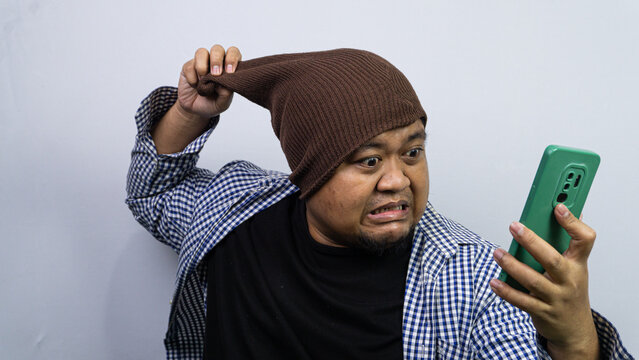 An angry adult Asian male forcefully pulls a brown knit beanie while glaring at a green smartphone. The frustrated individual exhibits a grimacing facial expression against a simple