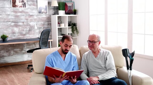 male caregiver reads from an open book with an older man seated on the sofa, representing companionship, engagement and supportive senior care.