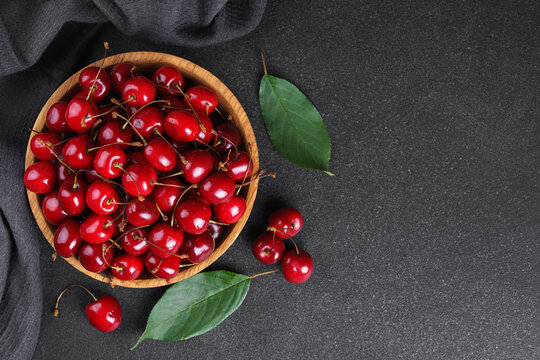 Fresh ripe sweet cherries in rustic wooden bowl with green leaves on black textured background. Top view flat lay with copy space. Juicy organic summer fruit, red berries, healthy seasonal food