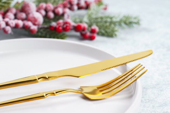 Festive holiday table setting with golden cutlery arranged on white plate with showy Christmas tree branch with red holly berry on light gray textured background. Knife and fork close up, macro