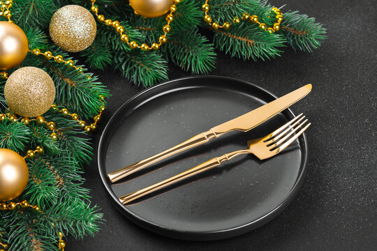 Elegant Christmas table setting with gold cutlery on a black plate, surrounded by evergreen branches, golden ornaments, beads and pinecones on a dark textured background