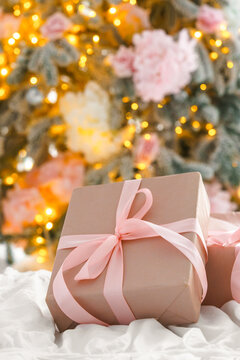 Beautifully wrapped Christmas present, gift box with soft pink ribbons lying on bed, with blurred festive lights and decorated tree in background, creating aesthetic, warm and elegant holiday mood.