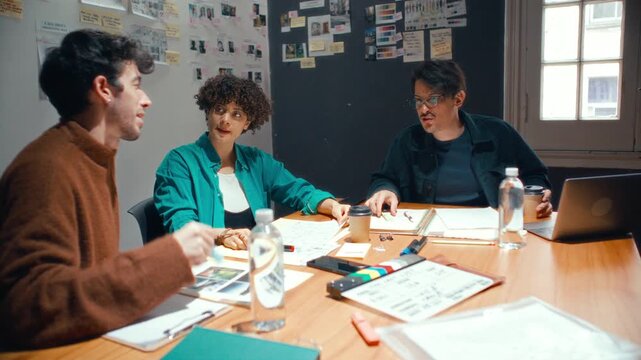Creative media team of three young professionals sitting around table, brainstorming ideas and reviewing references during film preproduction meeting in studio
