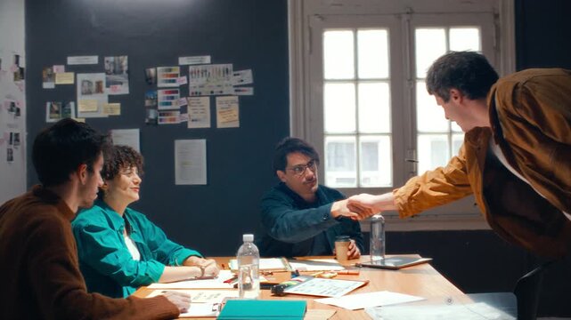 Team of professional filmmakers shaking hands and welcoming colleague, then discussing creative project during preproduction meeting in studio with reference moodboards in background