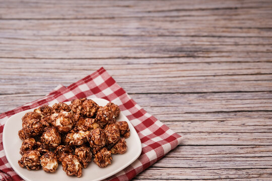 Crunchy chocolate caramel popcorn piled high on a white plate. Plate rests on a red-and-white checkered napkin. Wooden table and light gray paneling provide a cozy backdrop. Perfect for food blogs