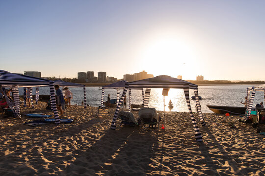 Beachgoers relaxing under the blue‑and‑white striped canopy tents are arranged along a sandy beach