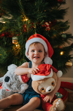 smiling blonde girl wearing santa hat hugs teddy bears by a Christmas tree