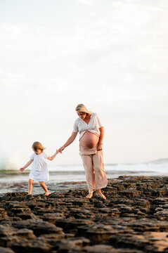 Expectant mother and daughter walking on rocky beach during sunset