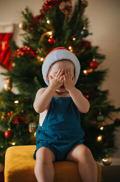 Mischievous blonde girl plays peekaboo wearing a Santa hat by a Christmas tree