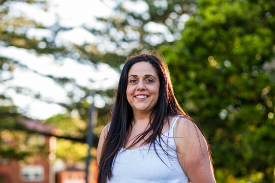 Woman stands in park with trees, smiling during sunset. She appears happy and relaxed.