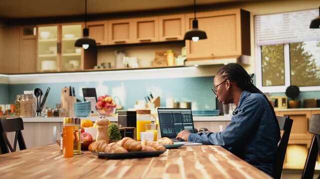 Digital nomad writes research data from charts while laptop displays analytics at kitchen table, combining task management and telecommuting productivity.