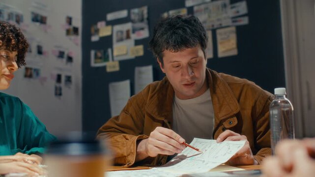 Сreative professional pointing to storyboard sketch with pencil, explaining script ideas to colleagues during preproduction meeting in studio with reference boards on walls